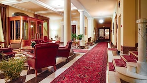 A hotel lobby with a checkered floor, red leather chairs, a large wooden cabinet, and a red carpeted corridor.
