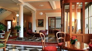 A hotel lobby with a checkered floor, red upholstered chairs, a wooden reception desk, and a large window.