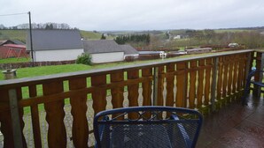 A wooden balcony with a metal chair, overlooking a rural landscape with houses and fields.