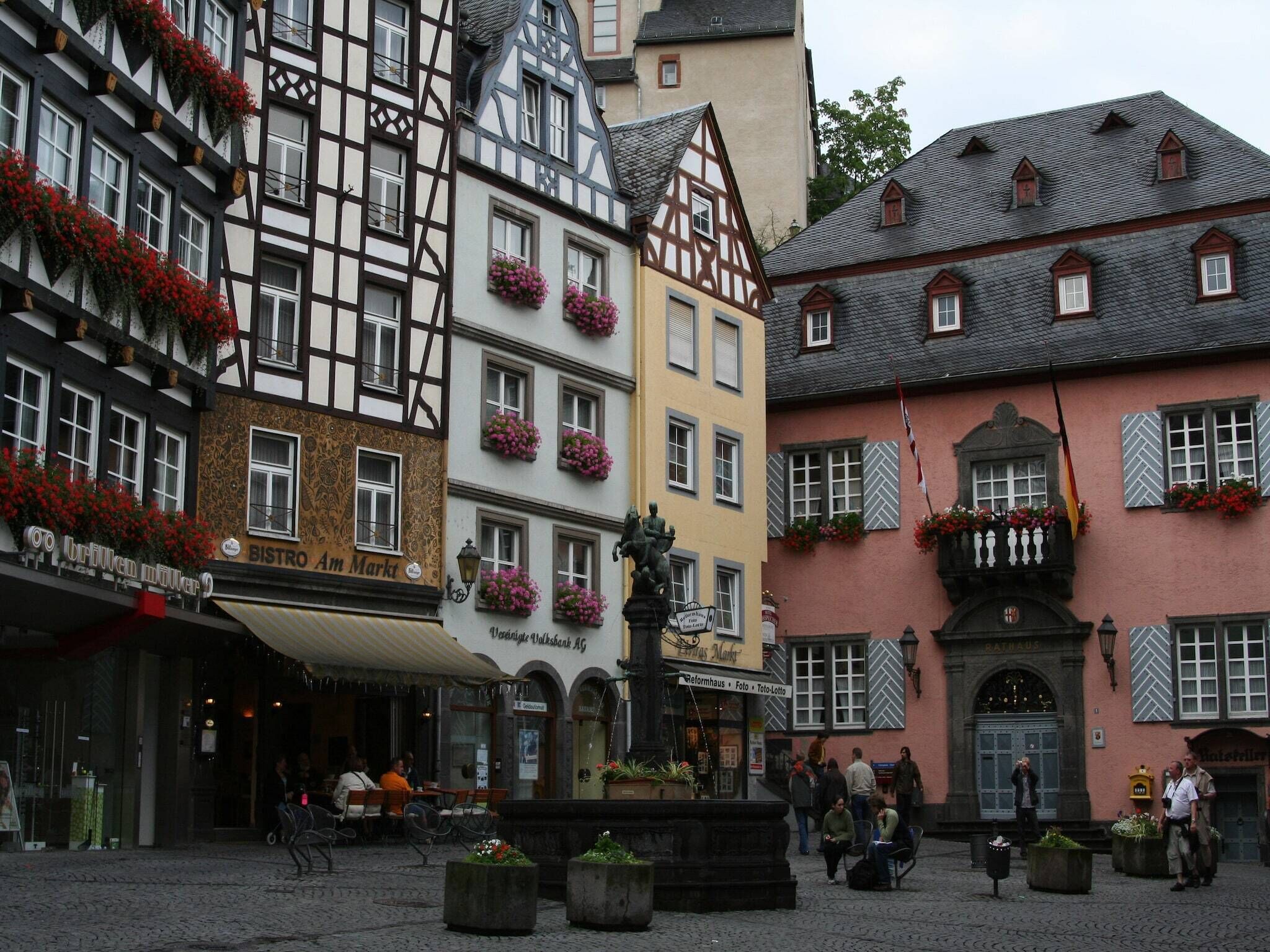 A traditional European street with historic buildings, a fountain, and outdoor seating areas.