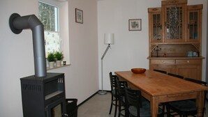 A kitchen with a wood-burning stove, a dining table with chairs, wooden cabinets, and a window with plants on the sill. - Bungalow near Eifel Ski Lift (Hinterhausen - Gerolstein)