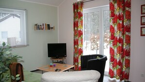 A living room with a television, a small table, a green chair, and a white chair. There is a window with blinds and a curtain with a floral pattern. - Bungalow near Eifel Ski Lift (Hinterhausen - Gerolstein)