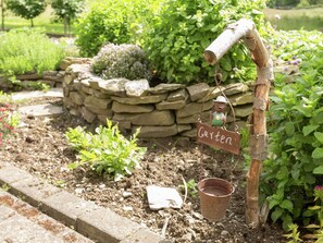 A rustic garden with a stone wall, a wooden sign reading "Garten", and a metal watering can hanging from a wooden post.