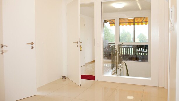 A room with a wooden ceiling, a glass door leading to a balcony with a railing and a chair, and a red mat at the door's base.