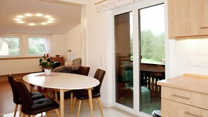 A modern kitchen with a large window, a dining area with a round table and chairs, and a sliding glass door leading to a balcony.