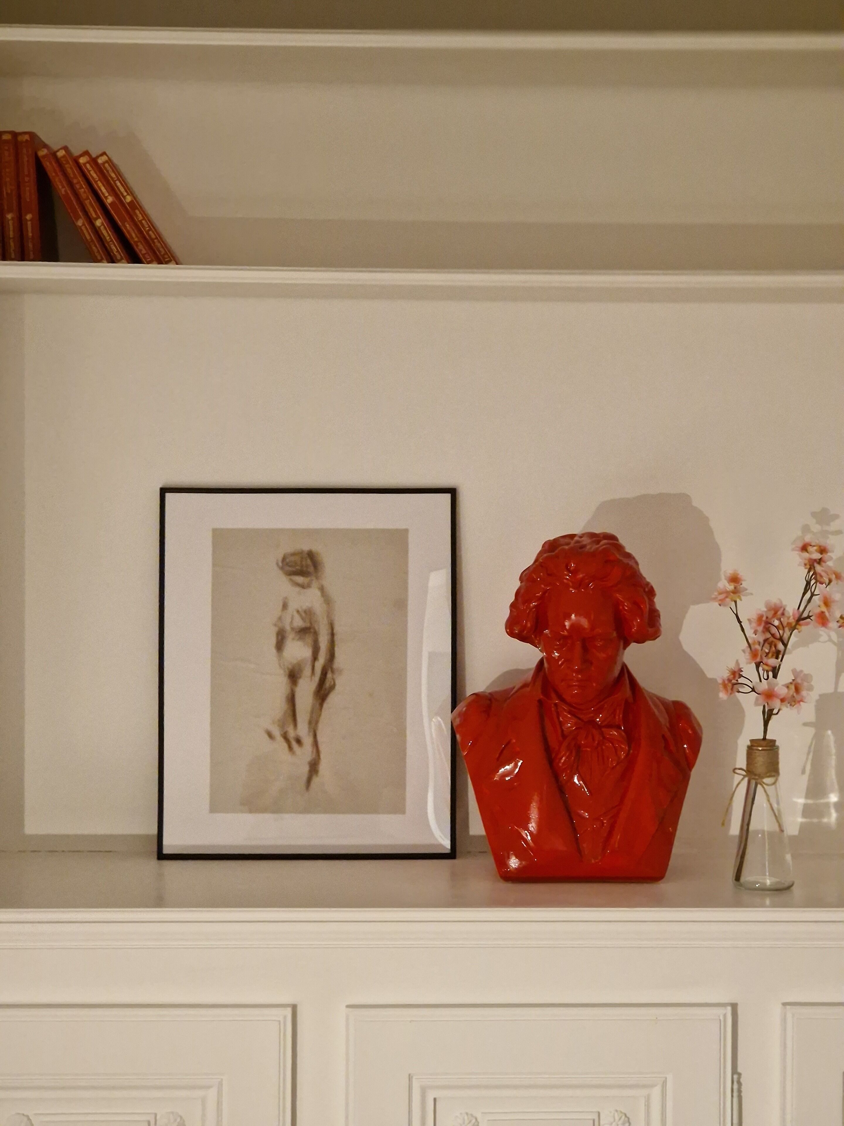 A white shelf with a framed black and white photograph, a red sculpture, and a small vase with flowers.