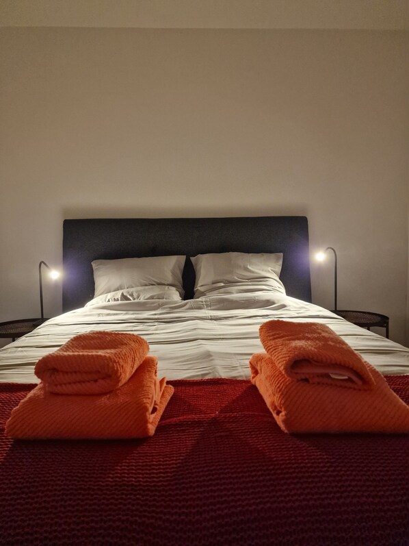 Cama bem arrumada com lençóis brancos e duas toalhas laranja dobradas sobre um cobertor vermelho.