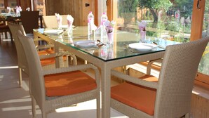 A dining area with wicker chairs and orange cushions, a glass-top table, and a view of greenery outside. - New York Houseboat (Srinagar)