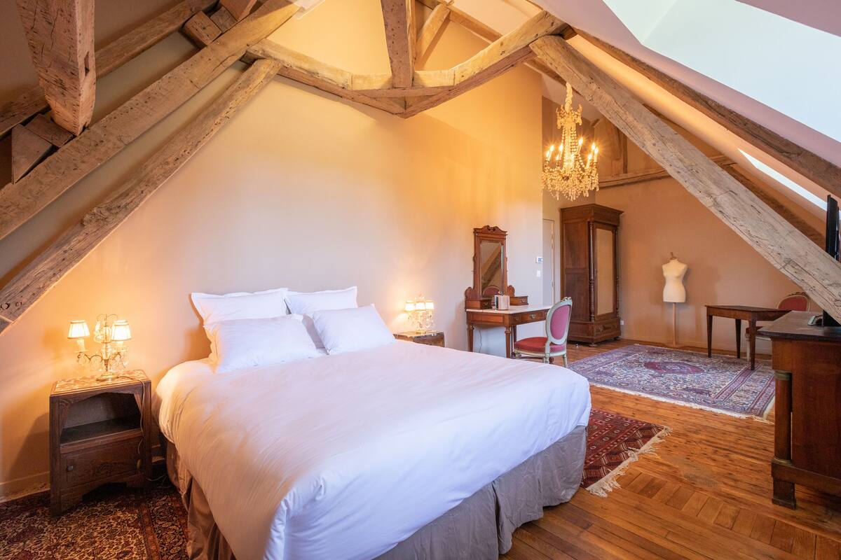 A bedroom with a large bed, wooden beams, a chandelier, a mirror, a dresser, and a rug.