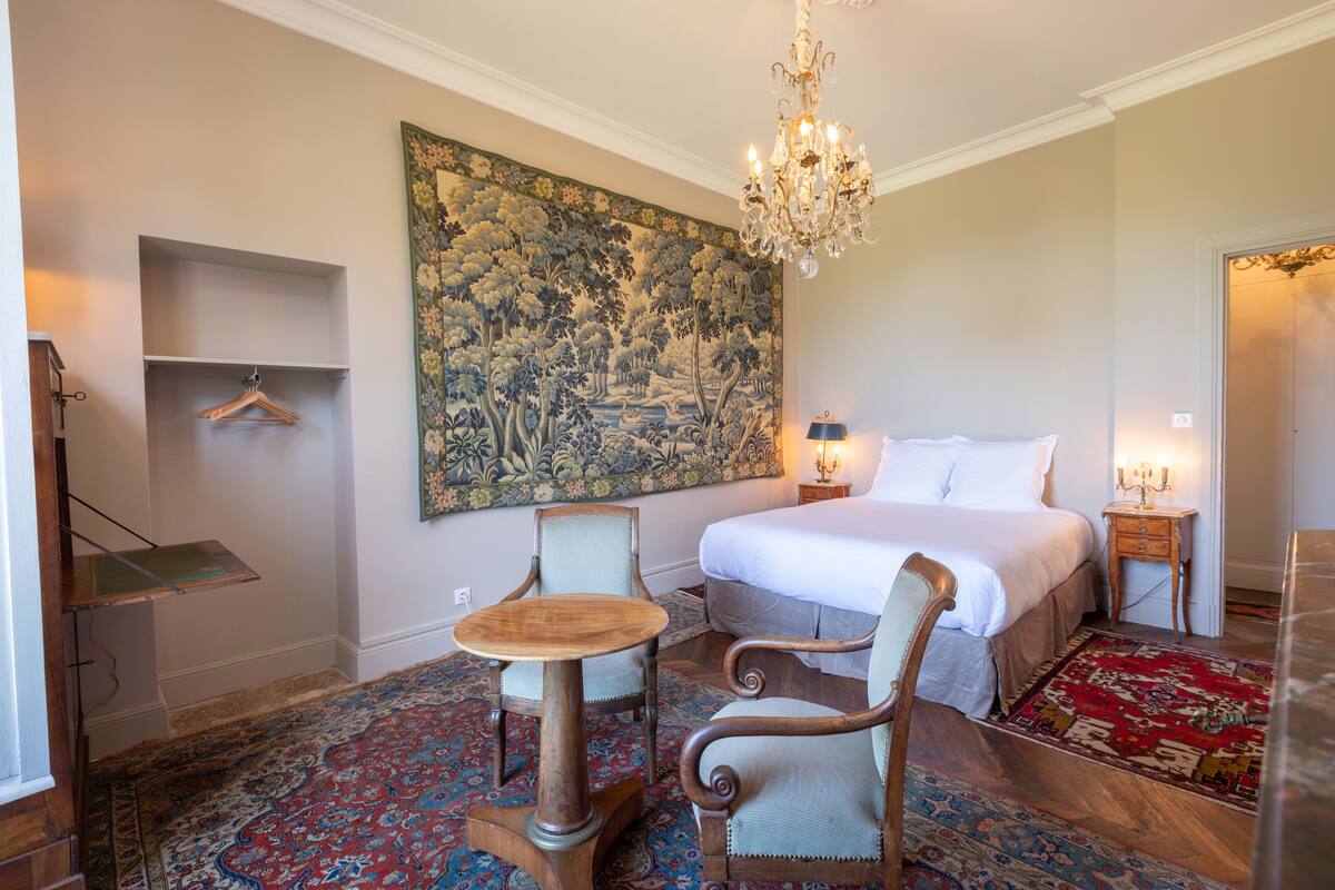A bedroom with a large bed, a chandelier, a wooden chair, a small table, a hanging coat rack, and a large floral tapestry on the wall.