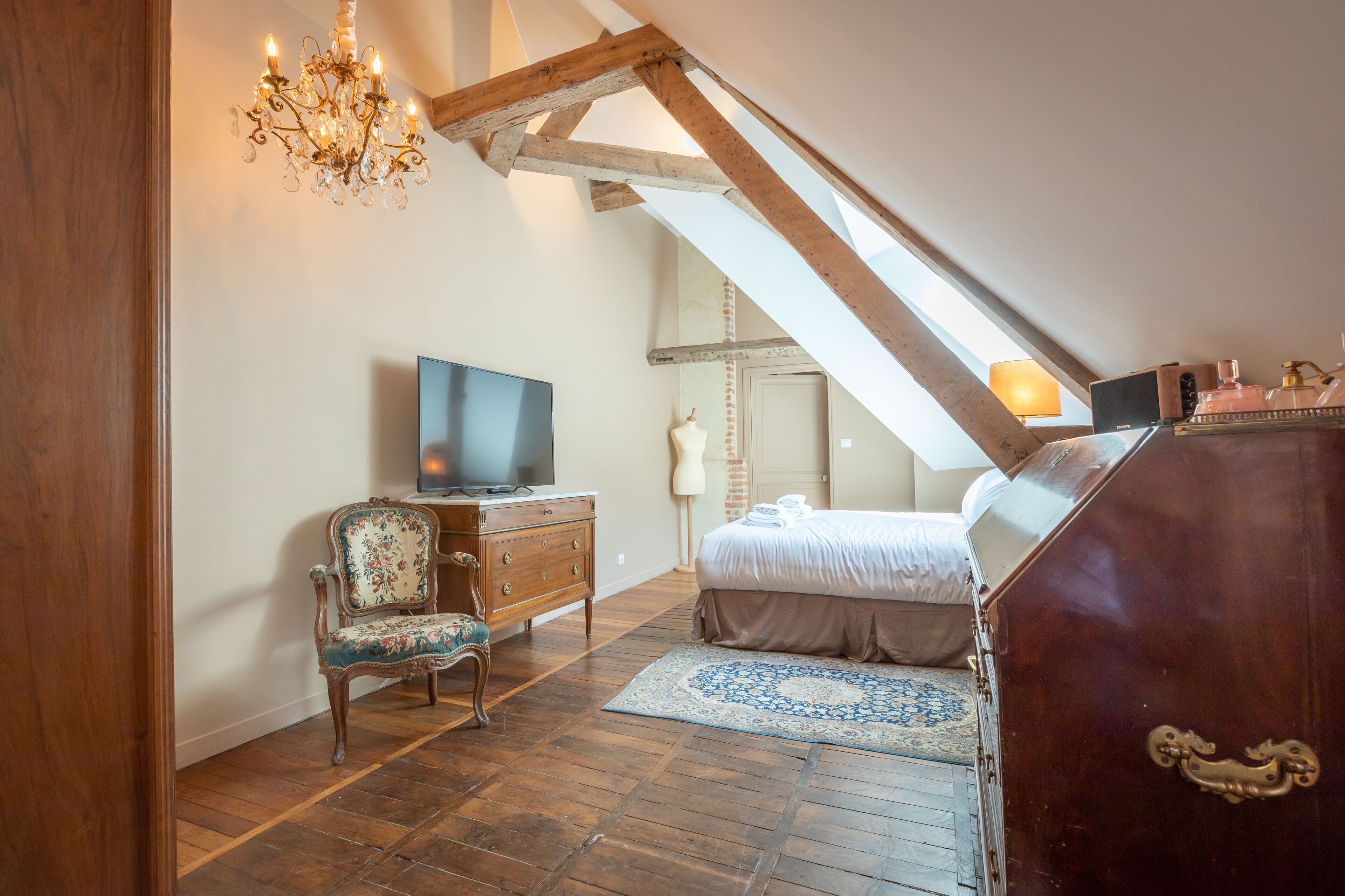 Een slaapkamer met een schuine plafond, een houten dressoir, een televisie, een stoel, een bed en een kroonluchter.