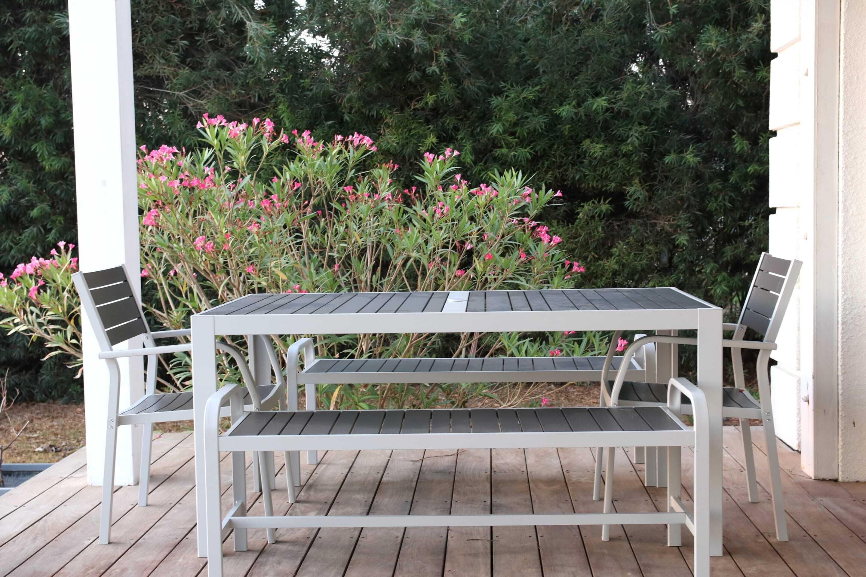 Ein weißer Tisch und Stühle im Freien auf einer Holzterrasse, im Hintergrund blühende, rosa Pflanzen.