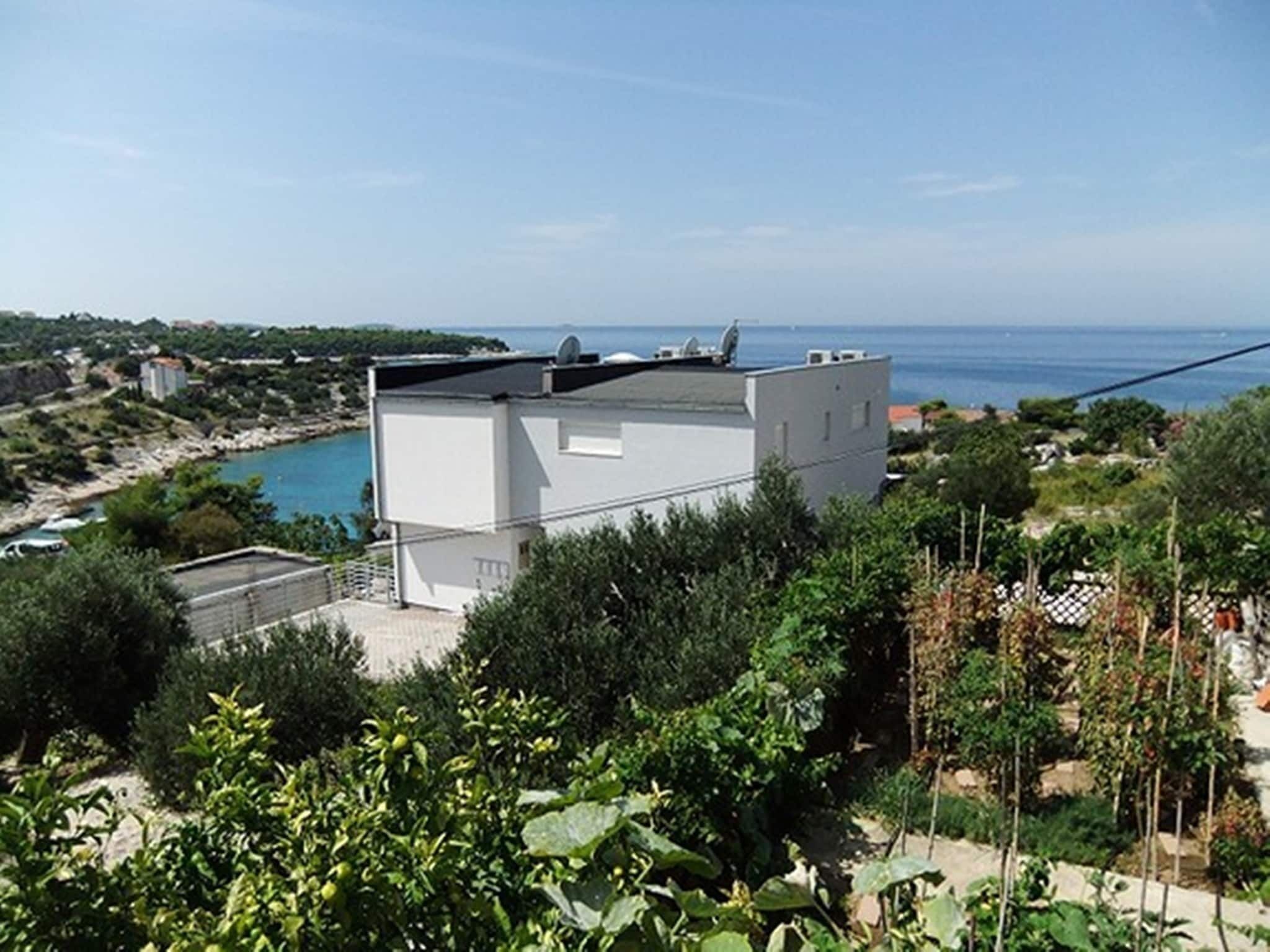 A modern building with a flat roof, surrounded by greenery and overlooking a coastal area.