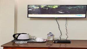 Un televisor montado en la pared, una cafetera, un vaso, una botella de agua y un control remoto sobre una mesa de madera.