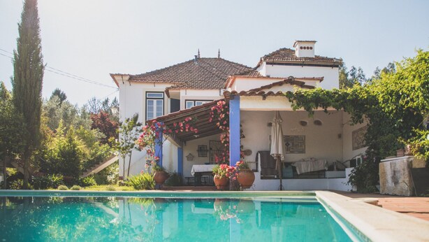 Vista a la piscina de una casa con porche cubierto, plantas en macetas y una piscina azul cristalina.