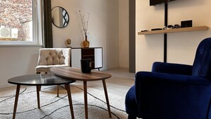 A modern living room with a blue armchair, a round coffee table, a sofa, a TV mounted on the wall, and a window with a view of a brick building. - La Little House de Dinant – Esc’Appart (Dinant)