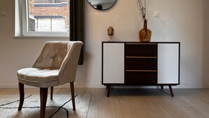 A modern living room with a tufted armchair, a dark wooden cabinet with drawers, a round mirror, and a vase with branches. - La Little House de Dinant – Esc’Appart (Dinant)