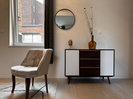 Una sala de estar moderna con un sillón capitoné, un mueble de madera oscura con cajones, un espejo redondo y un jarrón con ramas.