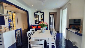 A modern kitchen with a central island, a dining area with a white table and chairs, and a living area with a sofa and colorful cushions. - In the blue painted blue (Campania)