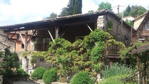 Un bâtiment rustique en pierre, doté d’un balcon en bois, entouré d’une végétation luxuriante et d’une cour recouverte de gravier.