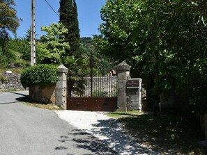 A gated entrance with a stone wall and metal gate, surrounded by lush greenery and a clear sky.