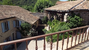 Un bâtiment traditionnel en pierre, avec un toit en tuiles, un balcon doté d’une balustrade en bois et une végétation luxuriante en arrière-plan.