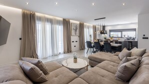 Ein modernes Wohnzimmer mit einem großen, beigefarbenen Ecksofa, einem runden Couchtisch und einem Essbereich, im Hintergrund ist eine Küche zu sehen.