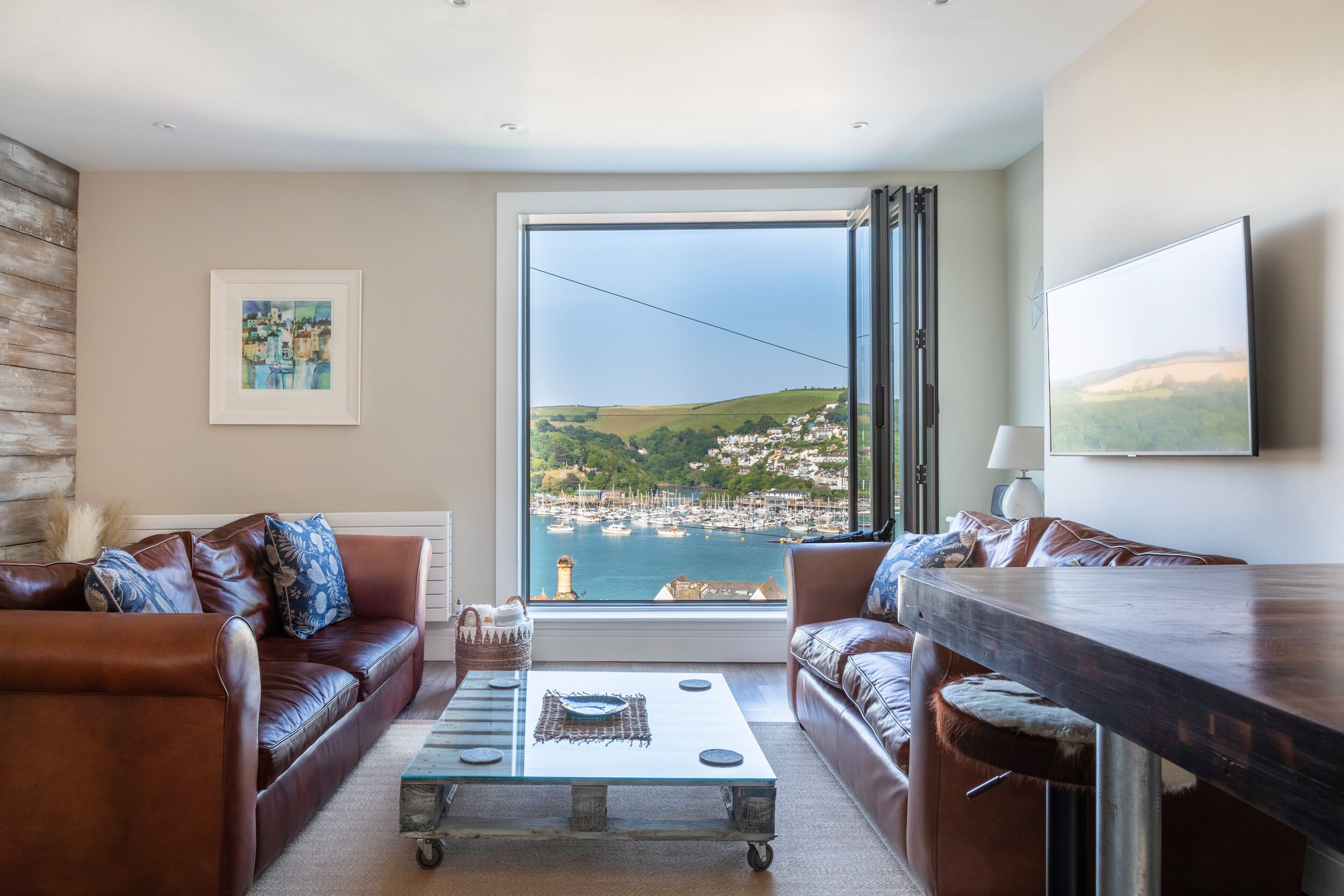A living room with a large window, a sofa, a coffee table, and a view of a coastal town.