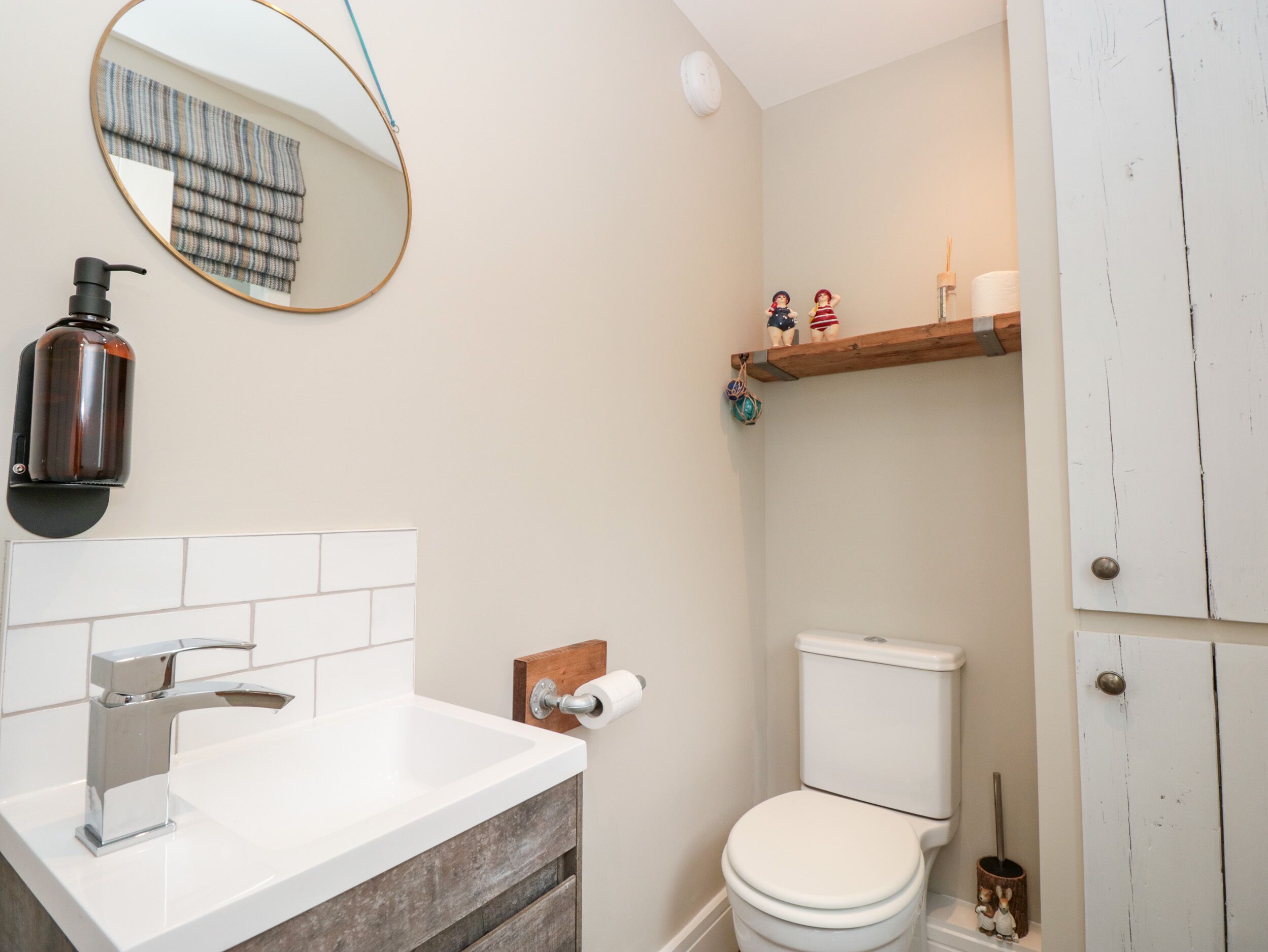 Et badeværelse med en hvid vask, kromarmatur og et rundt spejl. Der er et toilet med hvid låg og en lille hylde over det.