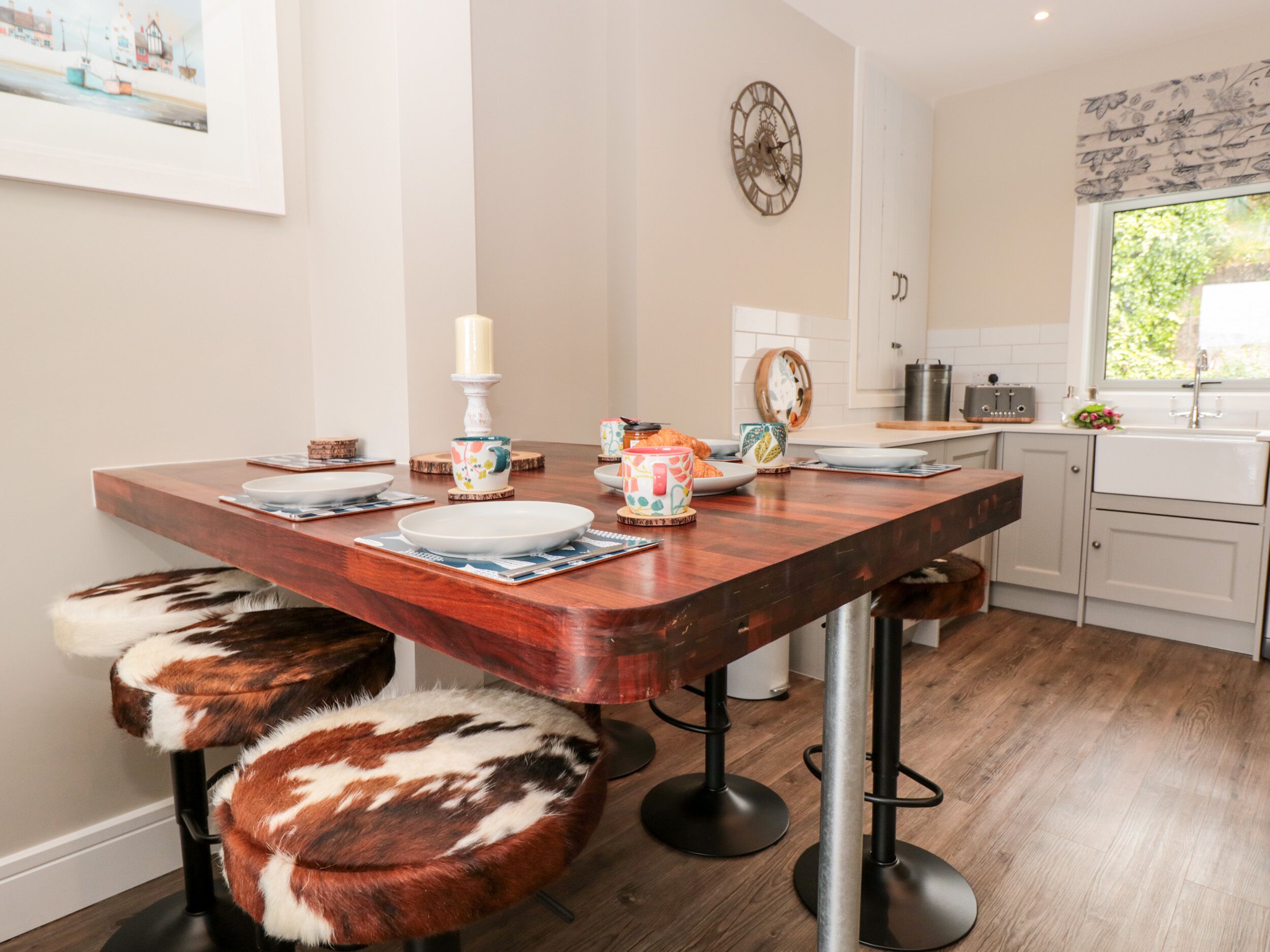 A modern kitchen with a wooden dining table, bar stools, and a window with a view of greenery.