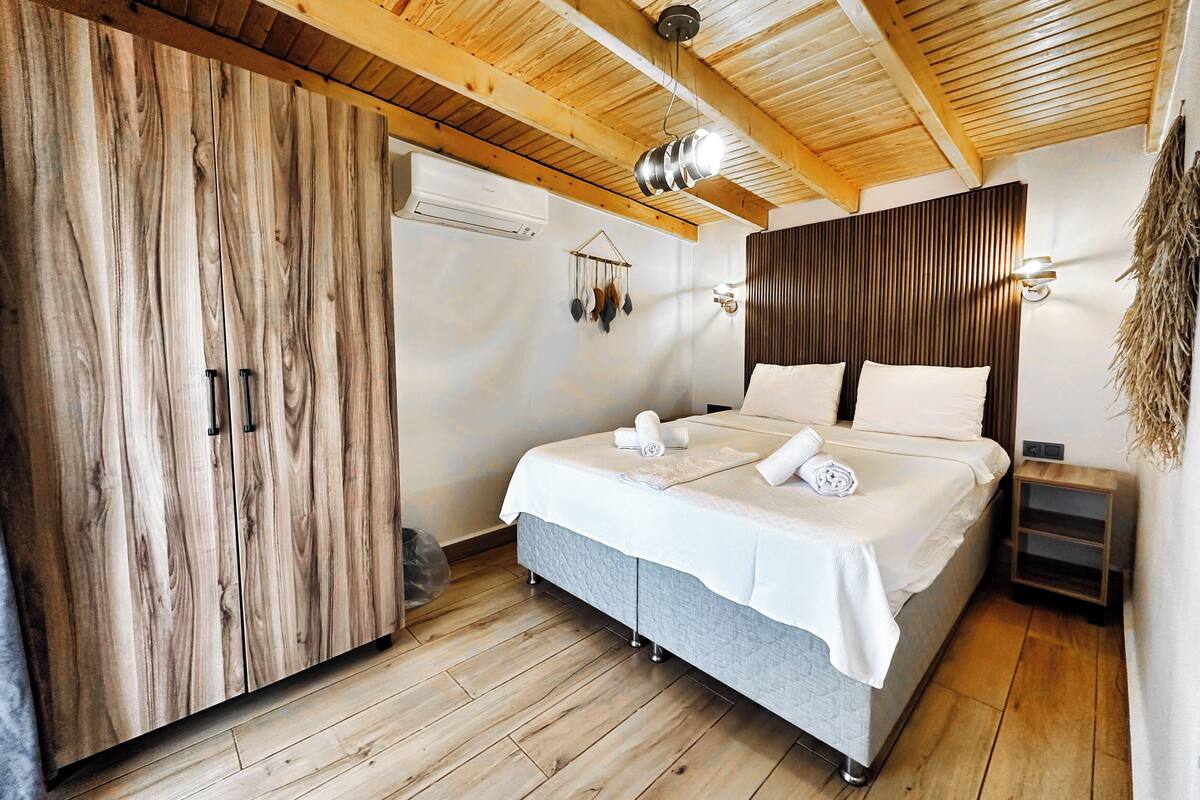 A bedroom with a wooden ceiling, a bed with white linens, a wooden wardrobe, and a wall-mounted light fixture.