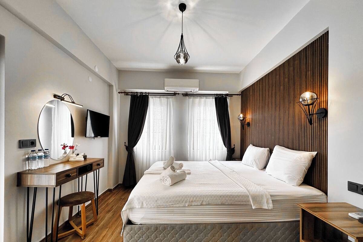 A modern hotel room with a large bed, a desk with a mirror, a television, and a wall-mounted light fixture.