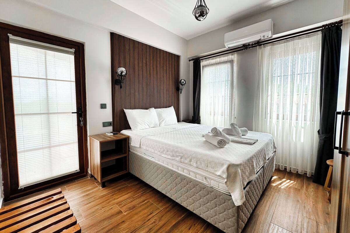 A modern hotel room with a large bed, wooden flooring, a nightstand, a wall-mounted lamp, and an air conditioning unit.