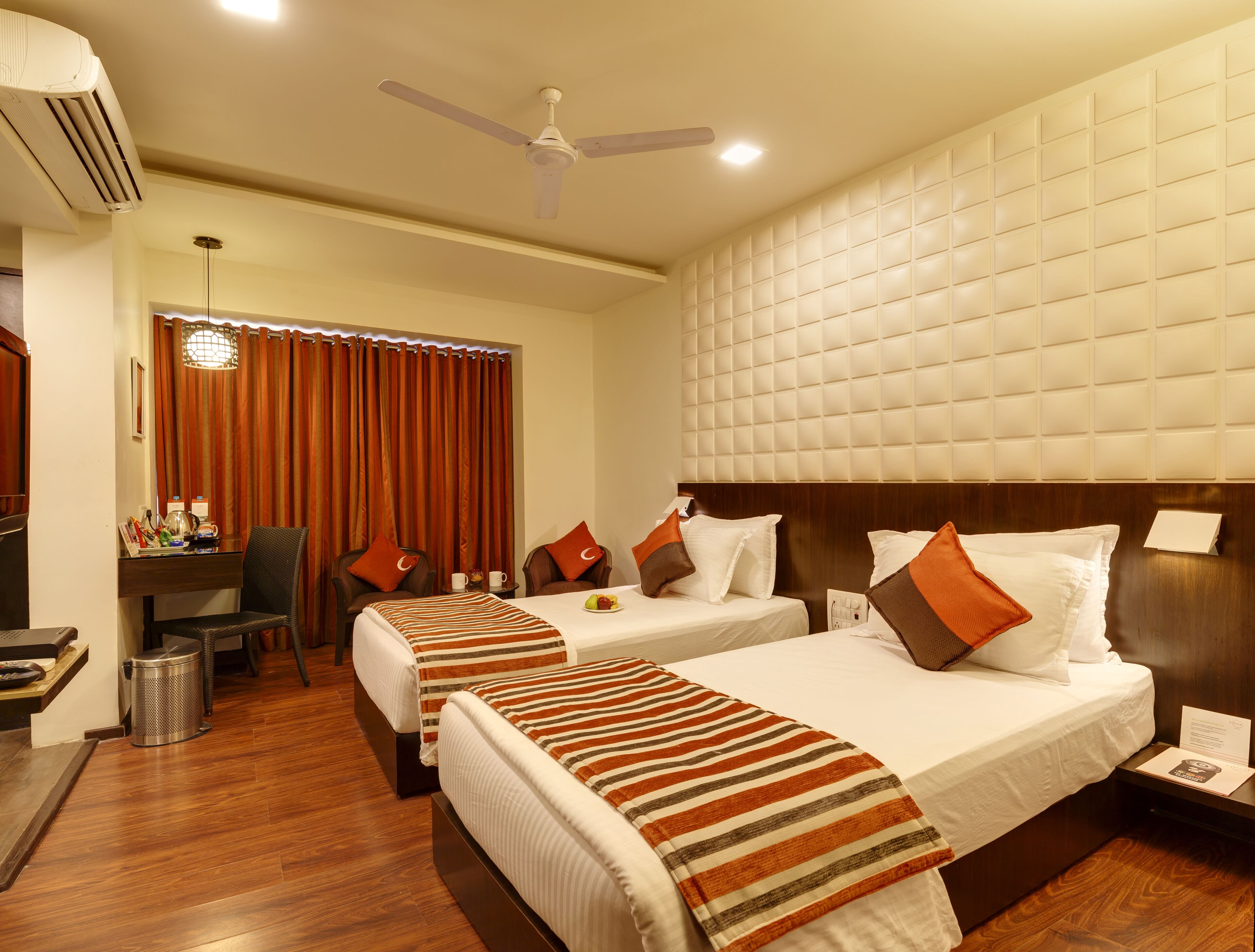 Quarto de hotel com duas camas, uma escrivaninha, cadeiras, um cesto de lixo e um ventilador de teto.