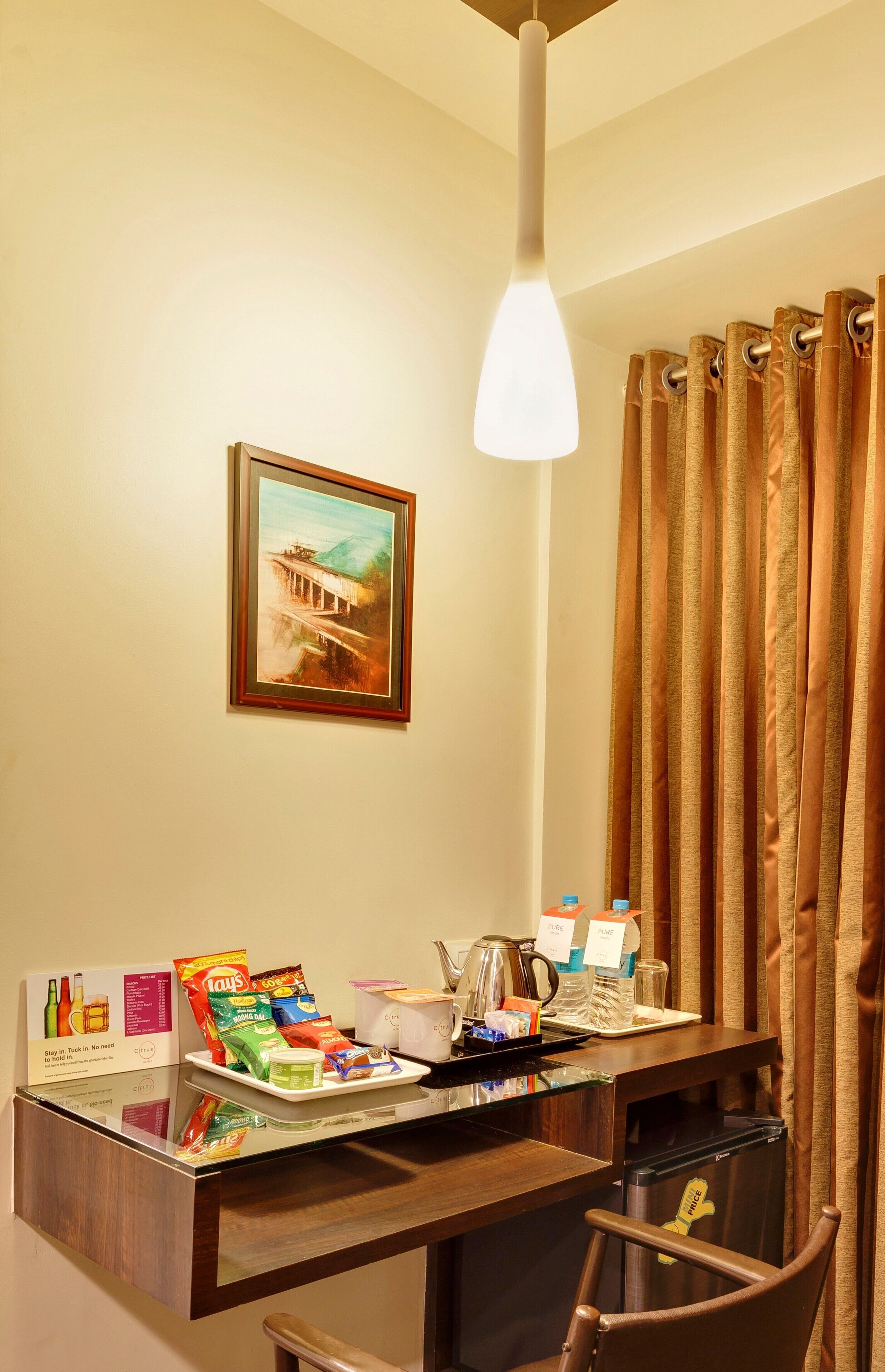 A hotel room with a wooden desk, a framed painting, a hanging light fixture, and a shower curtain.