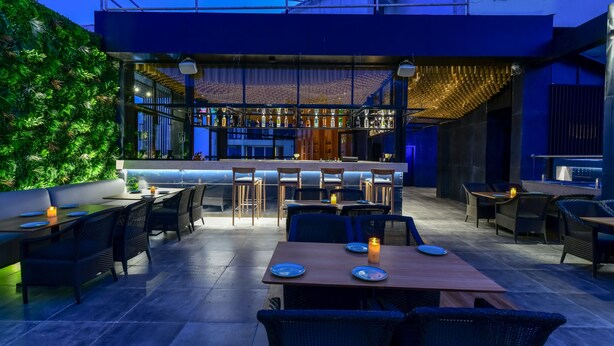 An outdoor dining area with tables and chairs, a bar in the background, and a green wall. - Regenta Place Raysons Kolhapur  (Kolhapur)