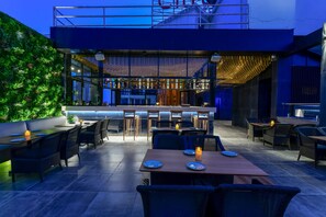 An outdoor dining area with tables and chairs, a bar in the background, and a green wall. - Regenta Place Raysons Kolhapur  (Kolhapur)