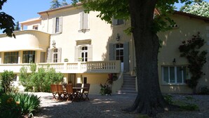 A two-story building with a balcony, a dining area with outdoor furniture, and a large tree in the foreground. - Country House in Provence with Garden Access (Valréas)