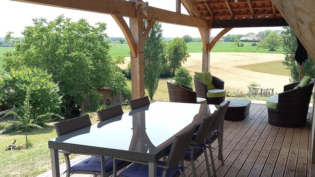 Une terrasse extérieure couverte, dotée d’une table et de chaises, de poutres en bois et offrant une vue sur un paysage verdoyant.