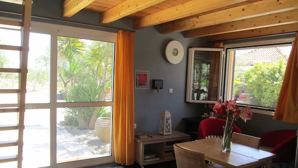 A room with a large sliding glass door leading to an outdoor area, a wooden ceiling, a small table with chairs, and a window with curtains. - Chalet near the beach in Messinia (Messinia)