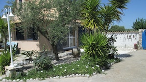 A building with a stone facade, a window, and a door. There are plants and a white wall with a blue door. - Chalet near the beach in Messinia (Messinia)
