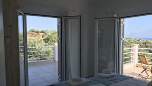 Une chambre avec deux lits, un balcon donnant sur un paysage verdoyant et la mer, et un fauteuil.