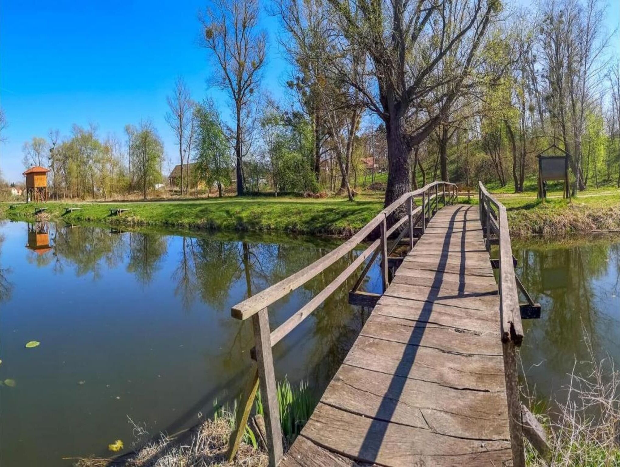 A wooden bridge over a calm pond with trees and a small structure on the left.