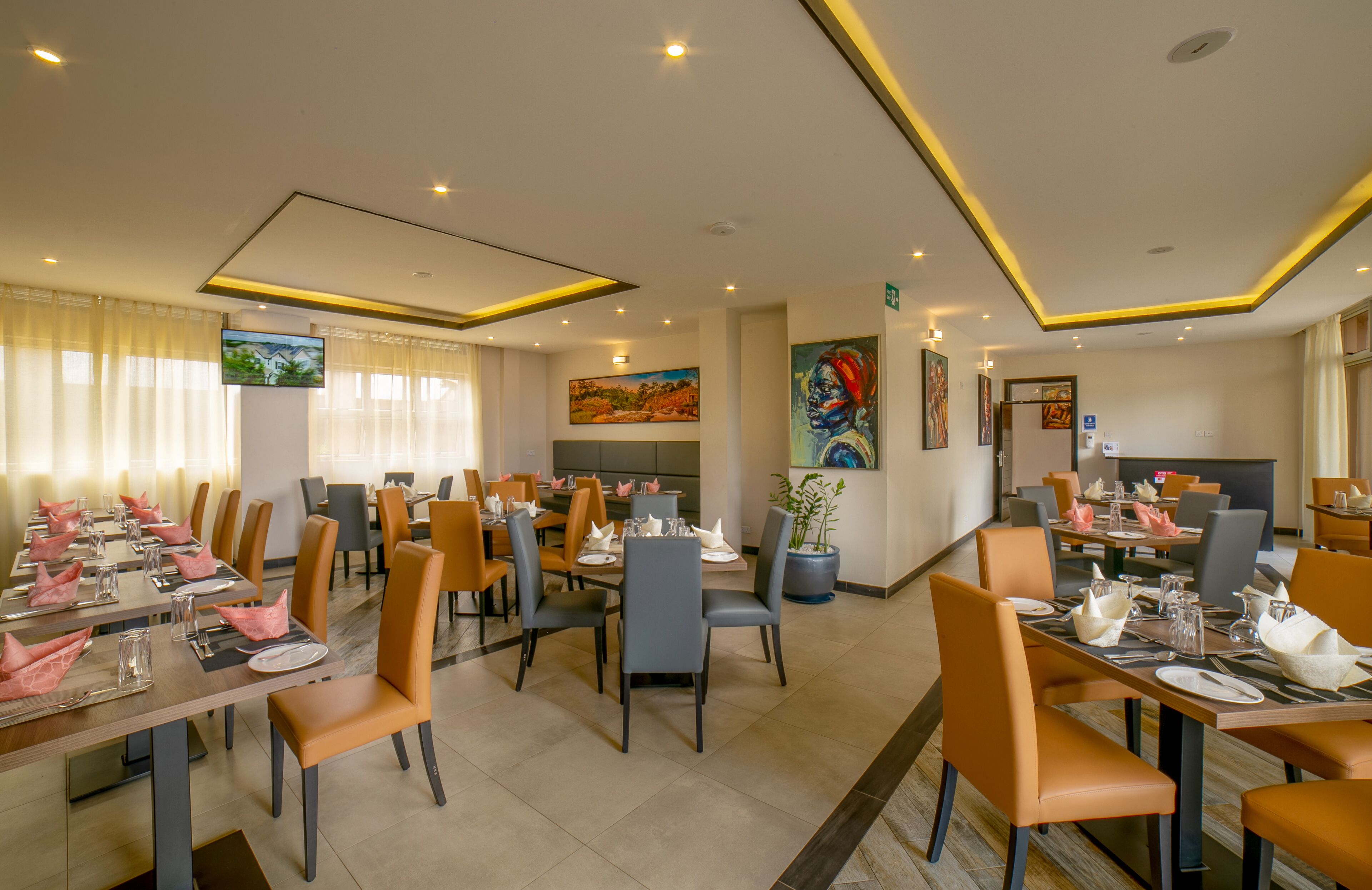 A hotel dining area with multiple tables set for guests, chairs, and a television screen on the wall.
