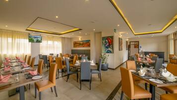 A hotel dining area with multiple tables set for guests, chairs, and a television screen on the wall.