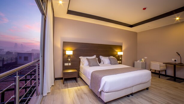 Quarto de hotel moderno com uma cama grande, abajures, uma escrivaninha e vista para a paisagem urbana.