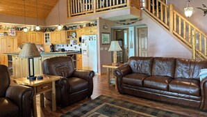 Ein geräumiges Wohnzimmer mit einem braunen Ledersofa, einem Holztisch und einem gemusterten Teppich. Eine Holztreppe mit Geländer führt auf eine obere Ebene.