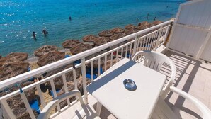 Un balcone con tavolo e sedie bianche affacciato su una spiaggia con ombrelloni di paglia e persone che fanno il bagno.