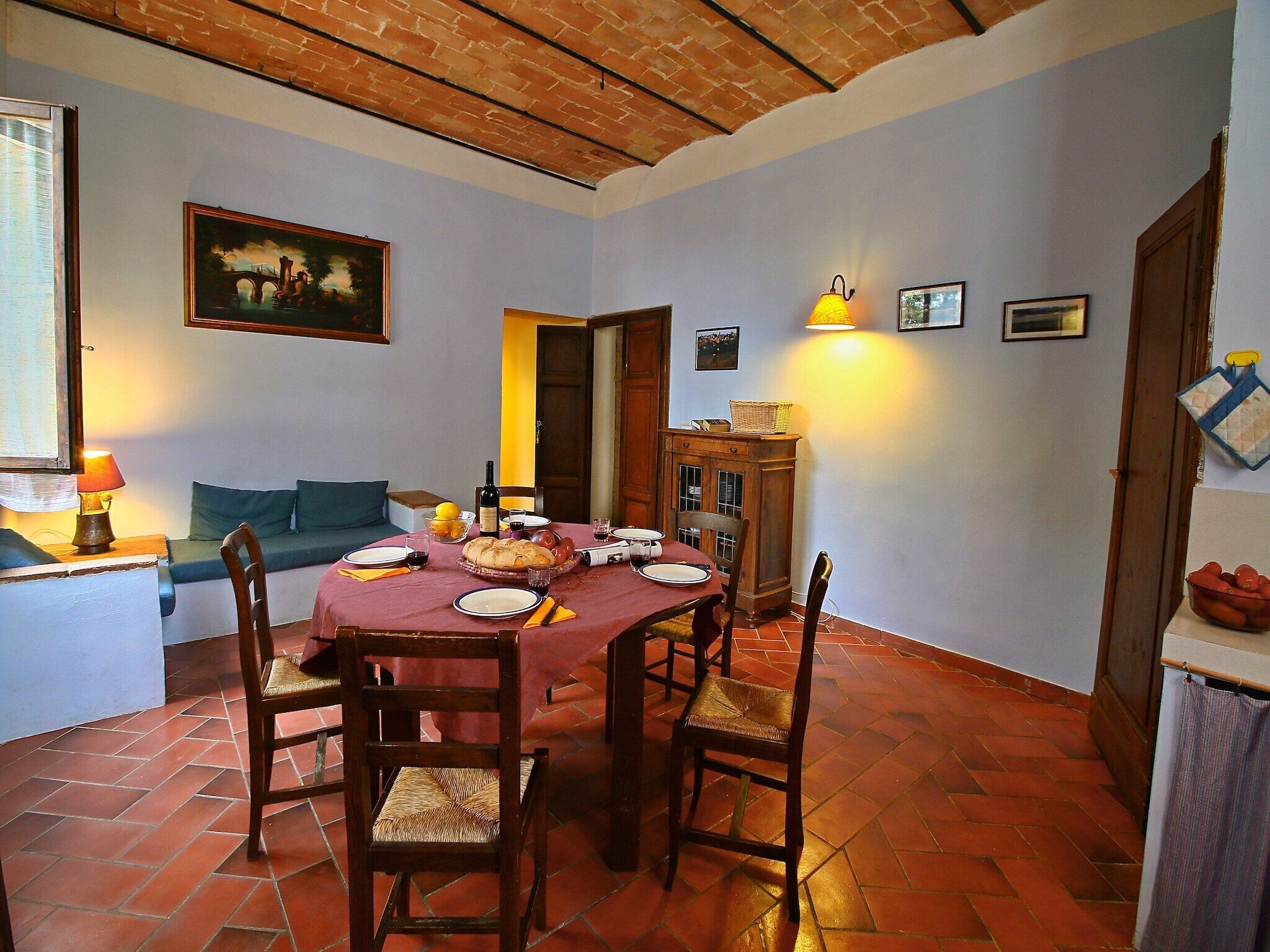 A dining area with a brick floor, wooden chairs, a red tablecloth-covered table, and a painting on the wall.