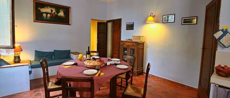 A dining area with a brick floor, wooden chairs, a red tablecloth-covered table, and a painting on the wall.